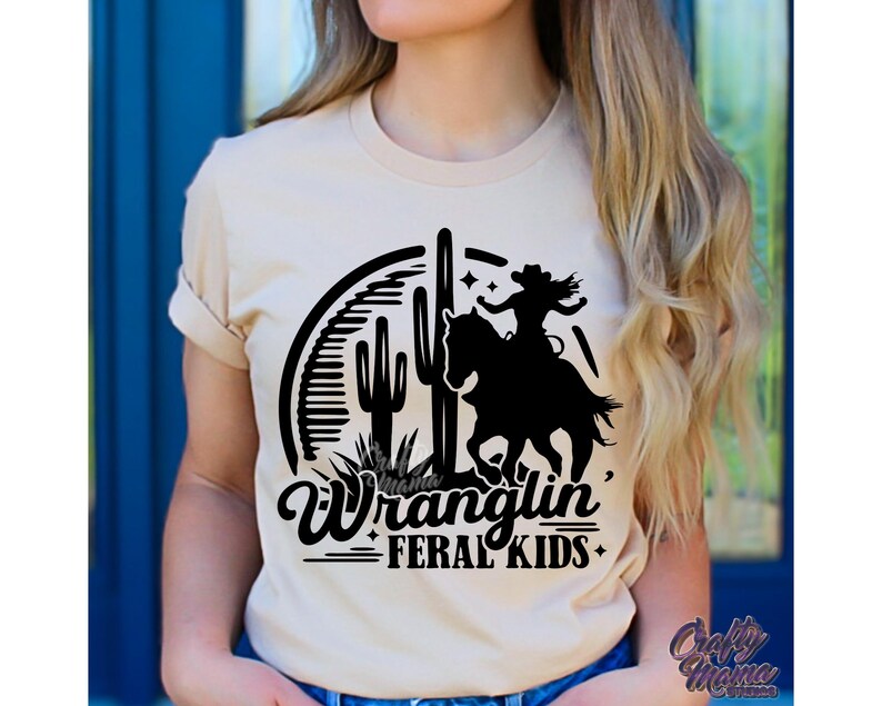 May include: Beige t-shirt featuring a black graphic of a person riding a horse, cacti, and the words "Wranglin' Feral Kids." The shirt has short sleeves and a crew neck.