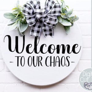 K&ouml;nnte beinhalten: Ein rundes, wei&szlig;es Holzschild mit einer schwarz-wei&szlig;en karierten Schleife und Gr&uuml;nzeug oben drauf. Das Schild sagt "Willkommen in unserem Chaos" in schwarzer, kursiv geschriebener Schrift.