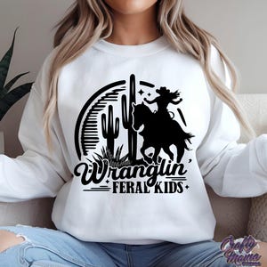 May include: White sweatshirt featuring a black graphic of a cowgirl riding a horse, cacti, and the words "Wranglin' Feral Kids." The design is enclosed in a semi-circle. The sweatshirt is a crew neck style.
