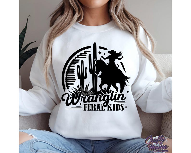 May include: White sweatshirt featuring a black graphic of a cowgirl riding a horse, cacti, and the words "Wranglin' Feral Kids." The design is enclosed in a semi-circle. The sweatshirt is a crew neck style.