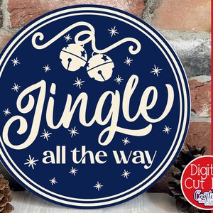 Op de afbeelding: Een rond blauw en wit bord met de tekst "Jingle all the way" in een wit cursief lettertype. Het bord is versierd met witte sneeuwvlokken en twee witte bellen.