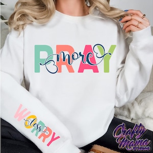 May include: White sweatshirt with the text "Pray More Worry Less" in rainbow colors. The text is in a stylized font with a heart shape in the middle of the word "Pray".