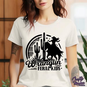 May include: Cream-colored t-shirt featuring a black graphic of a cowgirl riding a horse, cacti, and the text "Wranglin' Feral Kids." The design is set within a circular frame. The shirt has short sleeves.