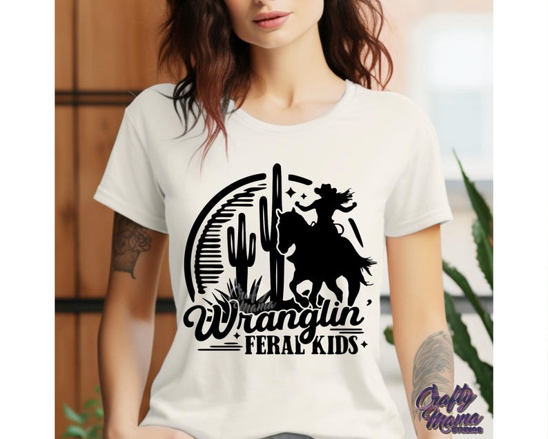 May include: Cream-colored t-shirt featuring a black graphic of a cowgirl riding a horse, cacti, and the text "Wranglin' Feral Kids." The design is set within a circular frame. The shirt has short sleeves.