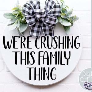 K&ouml;nnte beinhalten: Ein wei&szlig;es rundes Schild mit schwarzem Text, der "We're crushing this family thing" lautet. Das Schild ist mit einer schwarz-wei&szlig;en karierten Schleife und Gr&uuml;npflanzen verziert.