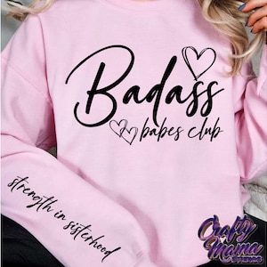 May include: Pink sweatshirt with the text "Badass babes club" in black cursive lettering with two hearts. The left sleeve has the text "strength in sisterhood" in black cursive lettering.
