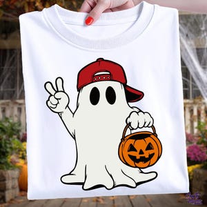 May include: White t-shirt featuring a cartoon ghost wearing a red baseball cap and holding an orange jack-o'-lantern bucket. The ghost is making a peace sign with its hand. The design is perfect for Halloween.