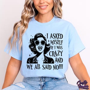 May include: A light blue t-shirt with a black and white graphic of a woman wearing sunglasses and the text "I asked myself if I was crazy and we all said no!!!"