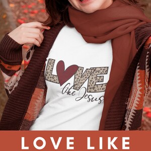 Puede incluir: Una camiseta blanca con un dise&ntilde;o gr&aacute;fico de la palabra "LOVE" en estampado de leopardo con un coraz&oacute;n rojo en el medio y las palabras "like Jesus" escritas debajo. La camiseta la lleva puesta una persona con una bufanda marr&oacute;n.