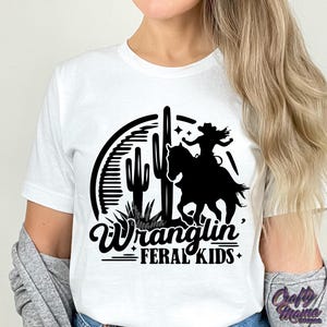 May include: White t-shirt with a black graphic of a cowgirl riding a horse, cacti, and the words "Wranglin' Feral Kids". The design is in a western style.