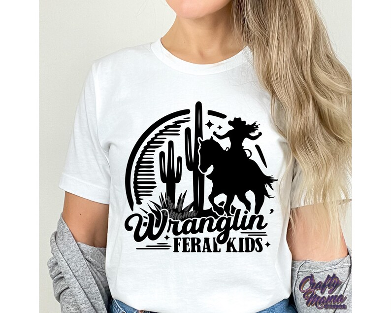 May include: White t-shirt with a black graphic of a cowgirl riding a horse, cacti, and the words "Wranglin' Feral Kids". The design is in a western style.