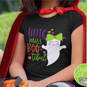 May include: A black t-shirt with a cute ghost graphic and the text "Little Miss Boo-tiful". The ghost has green and purple hair and a pink bow. The shirt is worn by a child wearing a red cape.