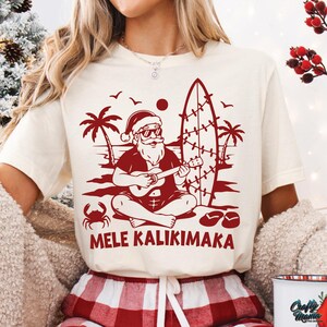 May include: Cream-colored t-shirt with a red graphic of Santa Claus playing a ukulele on a beach, with palm trees, a surfboard, and the text "MELE KALIKIMAKA".