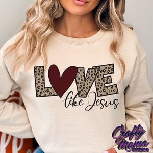 Puede incluir: Una sudadera beige con un dise&ntilde;o gr&aacute;fico de la palabra "LOVE" en estampado de leopardo con un coraz&oacute;n rojo en el centro. El texto "like Jesus" est&aacute; escrito debajo del gr&aacute;fico.