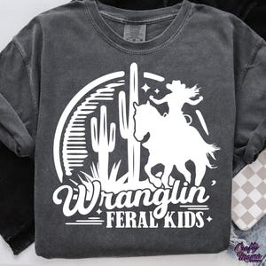 May include: Dark gray long-sleeve shirt with a white graphic. The graphic features a silhouette of a cowgirl riding a horse, a cactus, and the words "Wranglin' Feral Kids". The shirt has a crew neck and long sleeves.