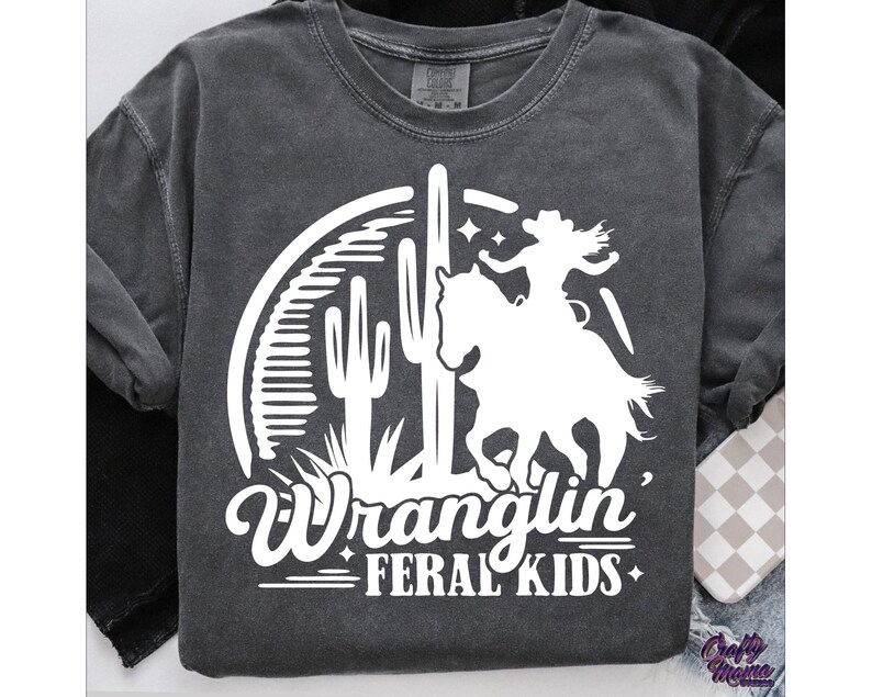 May include: Dark gray long-sleeve shirt with a white graphic. The graphic features a silhouette of a cowgirl riding a horse, a cactus, and the words "Wranglin' Feral Kids". The shirt has a crew neck and long sleeves.