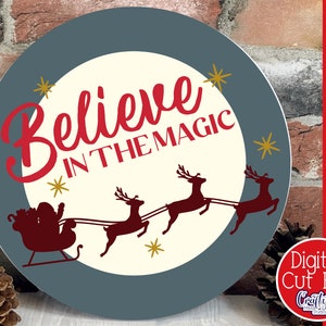 Op de afbeelding: Een rond, blauw en wit kerstbord met de tekst "Believe in the Magic" in rode cursieve letters. Het bord toont ook een silhouet van de kerstman in zijn slee getrokken door rendieren.