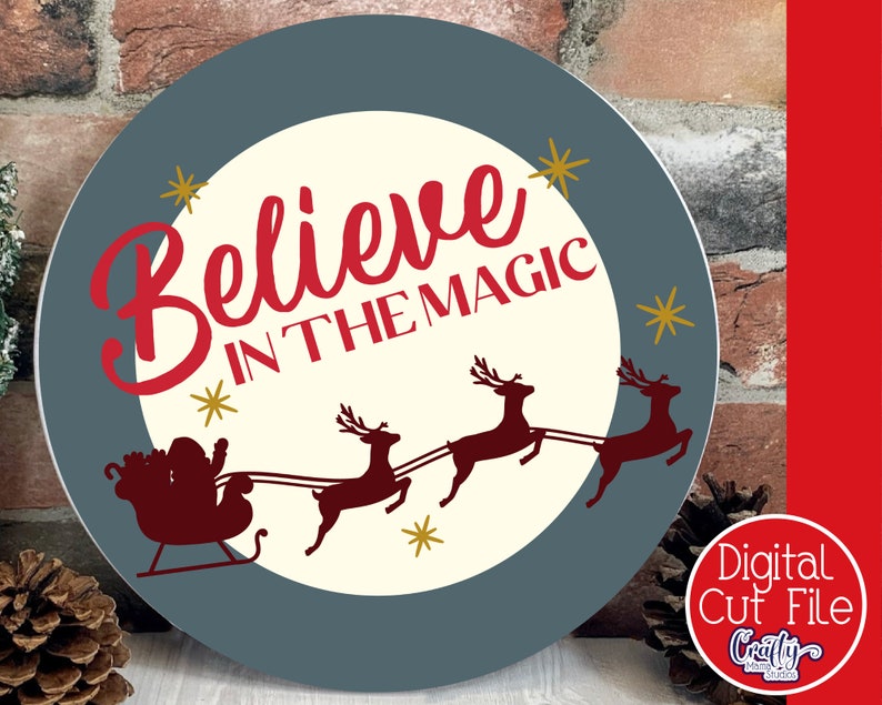 Op de afbeelding: Een rond, blauw en wit kerstbord met de tekst "Believe in the Magic" in rode cursieve letters. Het bord toont ook een silhouet van de kerstman in zijn slee getrokken door rendieren.