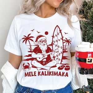 May include: White t-shirt with a red graphic of Santa Claus playing a ukulele on a beach, with a surfboard and palm trees. The text "MELE KALIKIMAKA" is printed below the graphic.