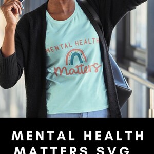 May include: A light blue t-shirt with the text "Mental Health Matters" and a rainbow graphic. The text is in a white font. The rainbow is made up of different colors, including red, orange, yellow, green, blue, indigo, and violet. The shirt is being worn by a person with a black cardigan and a black bag.