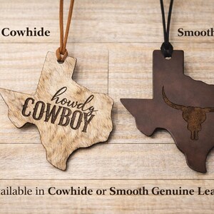 May include: Two Texas-shaped leather ornaments hanging from cords. The left ornament is tan cowhide with the text 'howdy COWBOY' engraved. The right ornament is dark brown smooth leather with an engraved longhorn skull graphic. Text labels read 'Cowhide', 'Smooth Leather', and 'Available in Cowhide or Smooth Genuine Leather'.