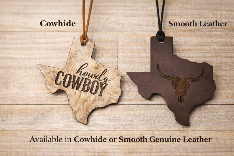 May include: Two Texas-shaped leather ornaments hanging from cords. The left ornament is tan cowhide with the text 'howdy COWBOY' engraved. The right ornament is dark brown smooth leather with an engraved longhorn skull graphic. Text labels read 'Cowhide', 'Smooth Leather', and 'Available in Cowhide or Smooth Genuine Leather'.