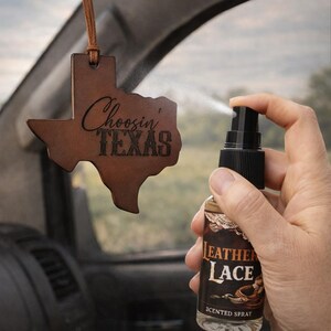 May include: A brown leather air freshener shaped like the state of Texas with the text Choosin' TEXAS, hanging from a brown cord. A clear spray bottle with a black nozzle and a label reading LEATHER LACE SCENTED SPRAY is being used to mist it.