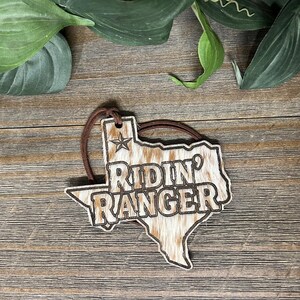 May include: A Texas-shaped ornament featuring a brown and white cowhide pattern with the text RIDIN' RANGER engraved in the center. A small star is positioned above the text, and a brown leather cord is attached to the top for hanging.