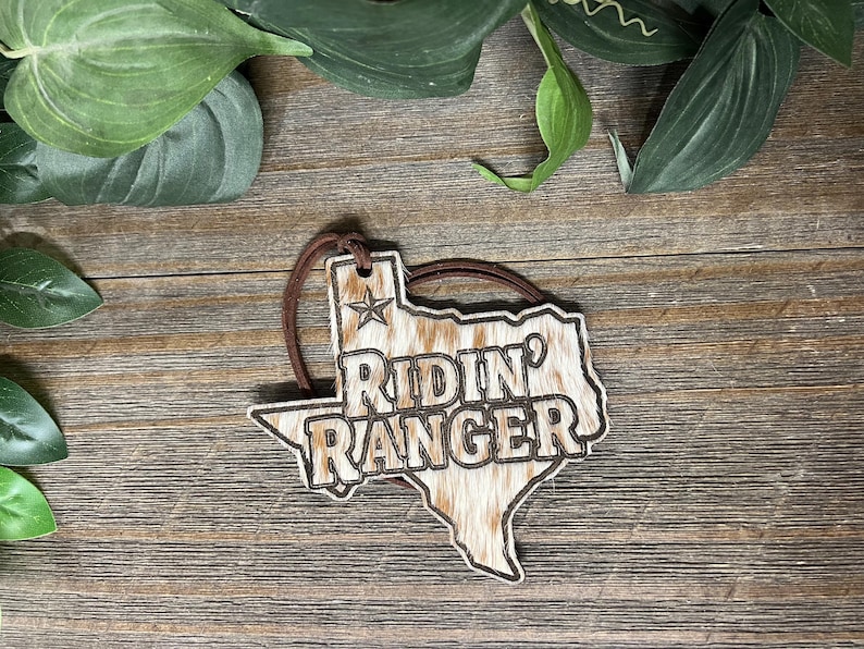 May include: A Texas-shaped ornament featuring a brown and white cowhide pattern with the text RIDIN' RANGER engraved in the center. A small star is positioned above the text, and a brown leather cord is attached to the top for hanging.