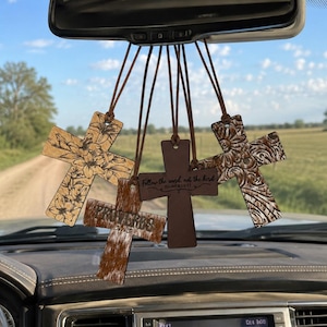 May include: A set of five hanging cross ornaments suspended from a rearview mirror. The crosses feature varied designs including floral patterns, a brown textured finish with the word PROVERBS, and a dark brown cross with the text Follow the herd not the bird.