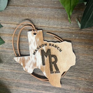 May include: A Texas-shaped ornament featuring a brown and white cowhide texture on the left side and a smooth tan finish on the right. It is engraved with the text Maxson Ranch, the letters MR, and EST 2021. A thin brown leather cord is attached.