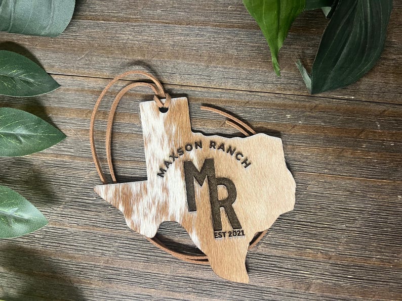 May include: A Texas-shaped ornament featuring a brown and white cowhide texture on the left side and a smooth tan finish on the right. It is engraved with the text Maxson Ranch, the letters MR, and EST 2021. A thin brown leather cord is attached.