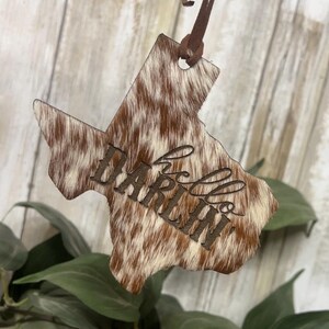 May include: A Texas-shaped ornament featuring a brown and white cowhide pattern with the word hello engraved in a cursive script across the center. The ornament is suspended by a thin brown leather cord attached to the top point of the shape.