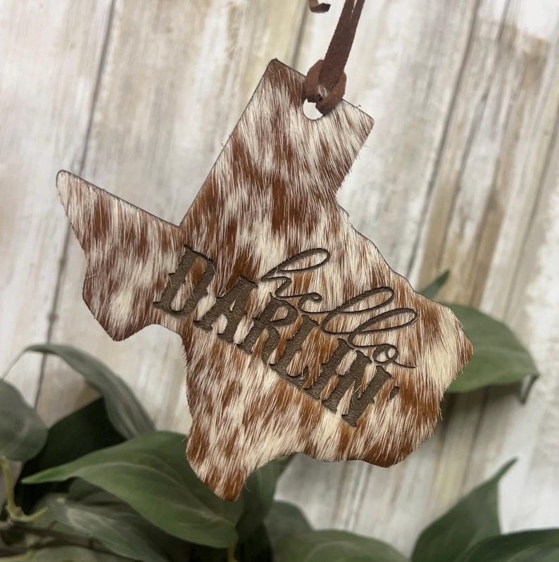 May include: A Texas-shaped ornament featuring a brown and white cowhide pattern with the word hello engraved in a cursive script across the center. The ornament is suspended by a thin brown leather cord attached to the top point of the shape.