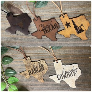 May include: Five Texas-shaped ornaments made of leather and hide, each featuring a leather cord for hanging. Designs include a longhorn skull, the text Choosin' TEXAS, an oil derrick with a star, the text hello DARLIN', and the text howdy COWBOY.