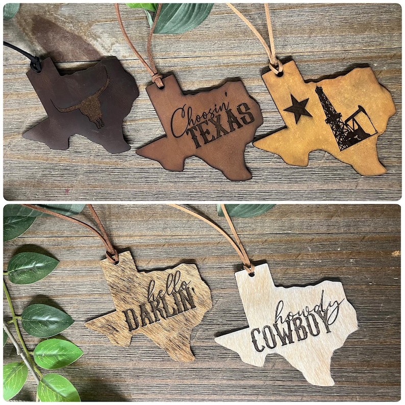 May include: Five Texas-shaped ornaments made of leather and hide, each featuring a leather cord for hanging. Designs include a longhorn skull, the text Choosin' TEXAS, an oil derrick with a star, the text hello DARLIN', and the text howdy COWBOY.