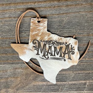 May include: A Texas-shaped ornament covered in brown and white cowhide with the word MAMA laser-engraved in the center. The ornament features a small hole at the top with a thin brown leather cord threaded through for hanging purposes.