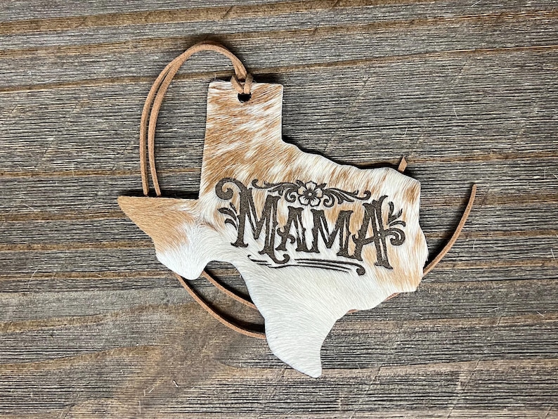 May include: A Texas-shaped ornament covered in brown and white cowhide with the word MAMA laser-engraved in the center. The ornament features a small hole at the top with a thin brown leather cord threaded through for hanging purposes.