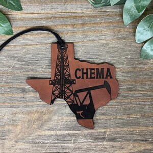 May include: A brown leather ornament shaped like the state of Texas, featuring a black laser-engraved design of an oil derrick and a pump jack. The word CHEMA is printed in black capital letters. A black cord is attached to the top for hanging.