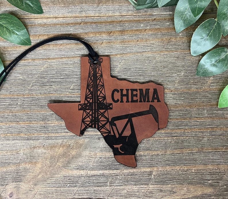 May include: A brown leather ornament shaped like the state of Texas, featuring a black laser-engraved design of an oil derrick and a pump jack. The word CHEMA is printed in black capital letters. A black cord is attached to the top for hanging.