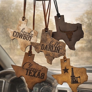 May include: Texas-shaped car air fresheners with various designs and phrases, including "Howdy Cowboy" and "Hello Darlin'". The air fresheners are made of wood and leather, with a rustic aesthetic. They hang from the rearview mirror.