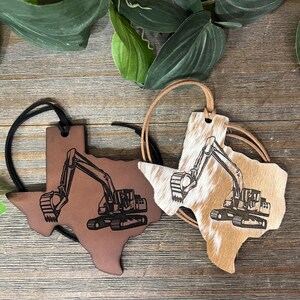 May include: Two Texas-shaped ornaments with black excavator engravings. One is smooth brown leather with a black cord, and the other is tan and white cowhide with a tan cord. Both feature a detailed construction vehicle design on the front surface.