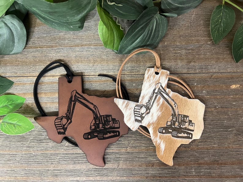 May include: Two Texas-shaped ornaments with black excavator engravings. One is smooth brown leather with a black cord, and the other is tan and white cowhide with a tan cord. Both feature a detailed construction vehicle design on the front surface.