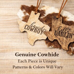 May include: Two Texas-shaped wooden ornaments with leather hanging cords. One light-colored ornament features the engraved text "howdy COWBOY" and the other darker, textured ornament features the engraved text "hello DARLIN'" in black.