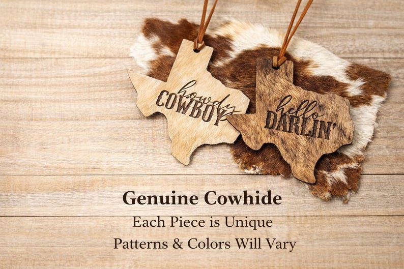 May include: Two Texas-shaped wooden ornaments with leather hanging cords. One light-colored ornament features the engraved text "howdy COWBOY" and the other darker, textured ornament features the engraved text "hello DARLIN'" in black.