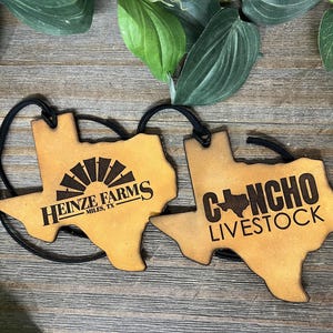 May include: Two tan leather-like Texas-shaped ornaments with black elastic cords. The left ornament features a windmill graphic and the text HEINZE FARMS MILES, TX. The right ornament displays the text CONCHO LIVESTOCK with a small Texas map icon.