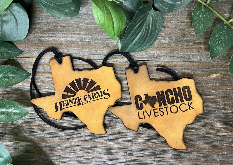May include: Two tan leather-like Texas-shaped ornaments with black elastic cords. The left ornament features a windmill graphic and the text HEINZE FARMS MILES, TX. The right ornament displays the text CONCHO LIVESTOCK with a small Texas map icon.