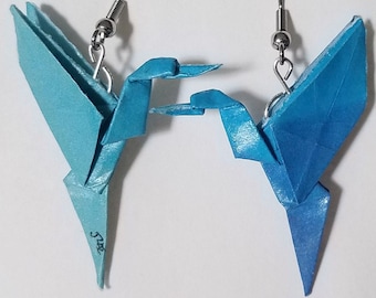 Può includere: Un paio di orecchini a forma di uccello origami blu con ganci argentati. Gli uccelli si guardano e hanno un gradiente di colori blu.