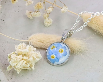 May include: A silver chain necklace with a round pendant featuring a cluster of blue forget-me-not flowers pressed inside a clear glass dome.
