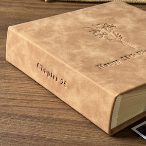 Peut inclure: Un album photo de couleur beige avec des d&eacute;tails floraux en relief et le texte "Chapter 25" et "Happy 25th Birthday". L'album a une couverture textur&eacute;e et un design classique.